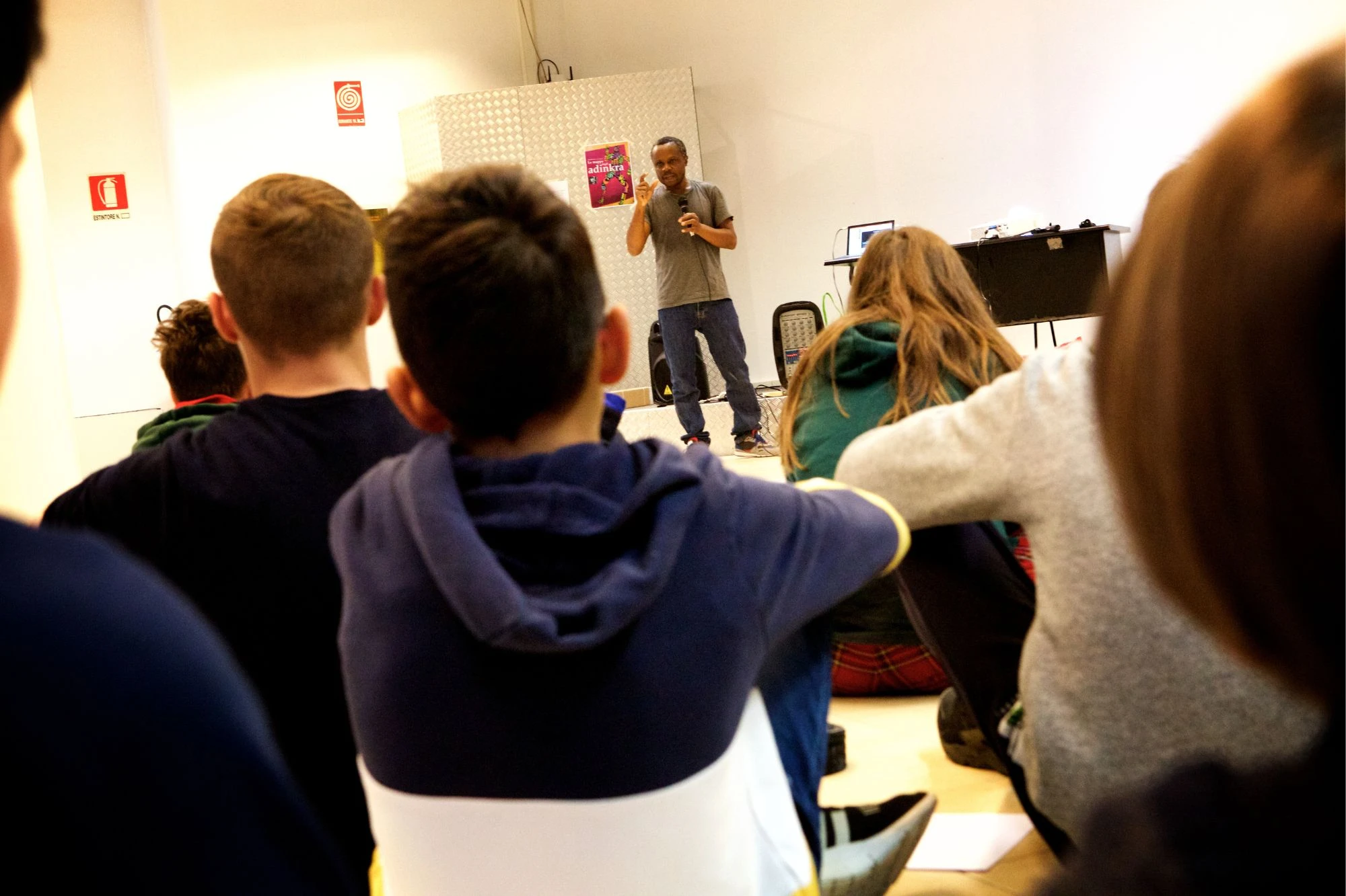 Henri Olama racconta i simboli Adinkra accanto al manifesto del libro, davanti a un gruppo seduto a terra ripreso di spalle